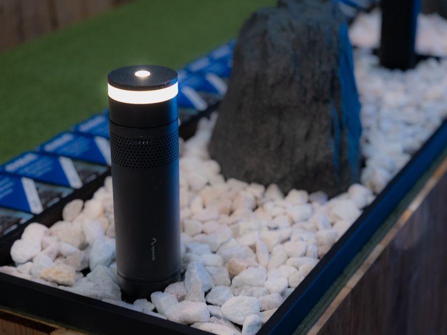 A lit bollard speaker in front of a rock speaker in a bed of white stones