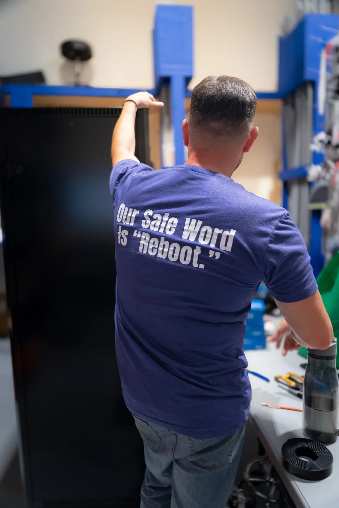 An AVAS team member from behind in a navy shirt that reads Our Safe Word Is Reboot, steadying a black equipment rack cabinet