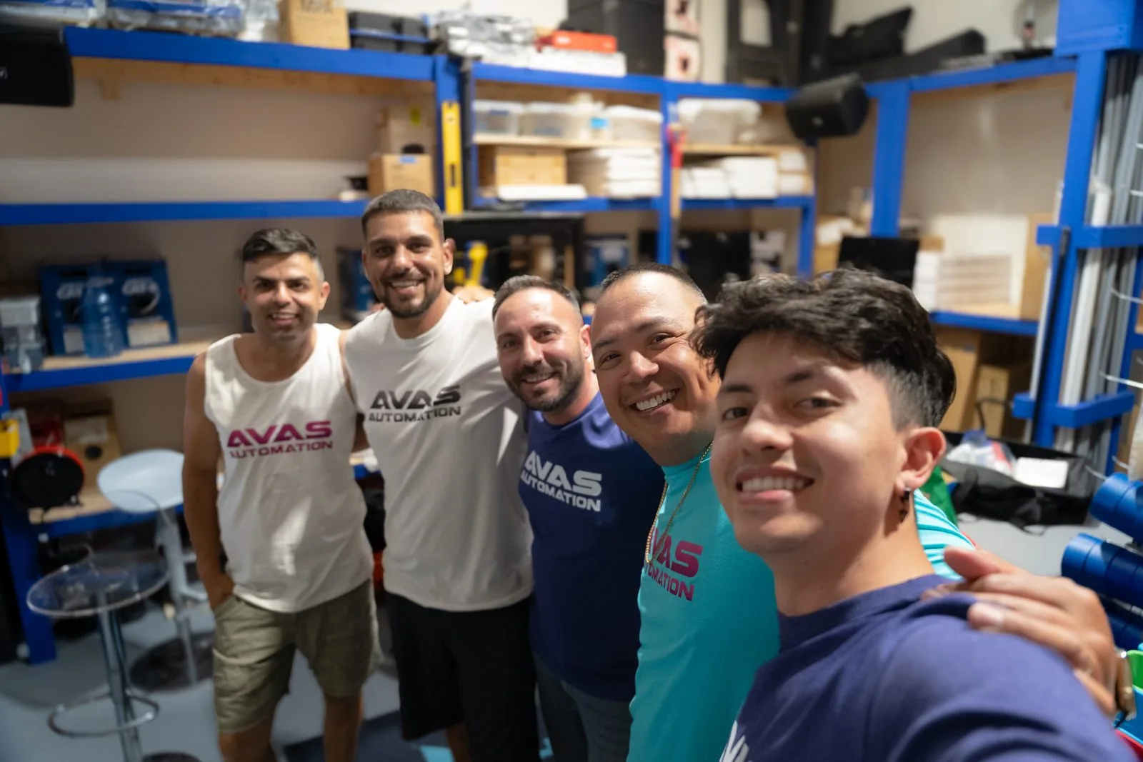 Five members of the AVAS Automation team smiling for a selfie in a workshop with blue shelving