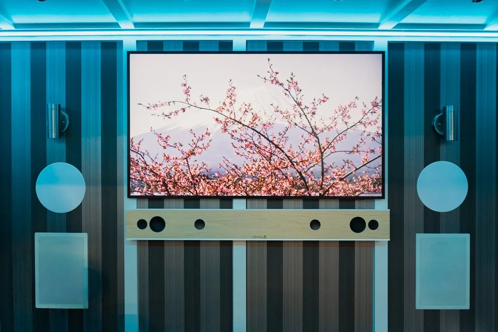 The lounge wall of the AVAS trailer with its coffered ceiling lit cool blue, a TV between two wall sconces and two round speakers, and a wood soundbar below